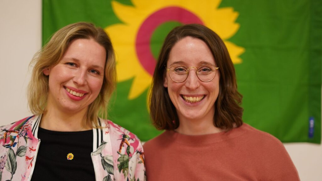 Doppelporträt Swantje Schendel mit Louise Bohne vor einer Fahne der grünen Partei (Die Sonnenblume darauf hat einen rosa Kreis.)