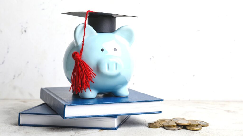 Ein blaues Porzelan-Schweinchen mit Doktorhut steht auf zwei Büchern, davor liegen diverse Euromünzen.