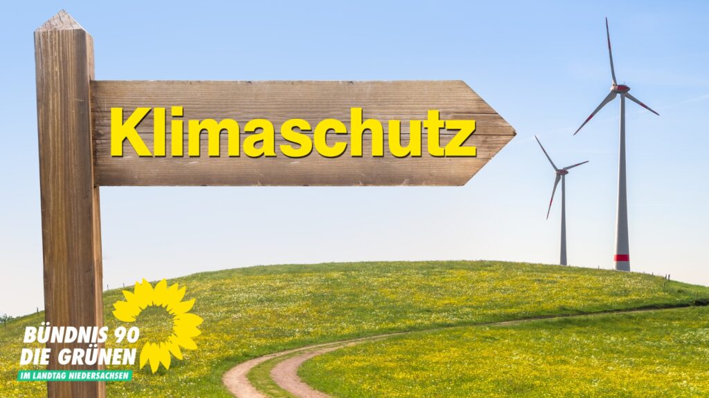 Auf einem großen hölzernen Pfeil steht "Klimaschutz" und er weist auf zwei Windräder im Hintergrund.