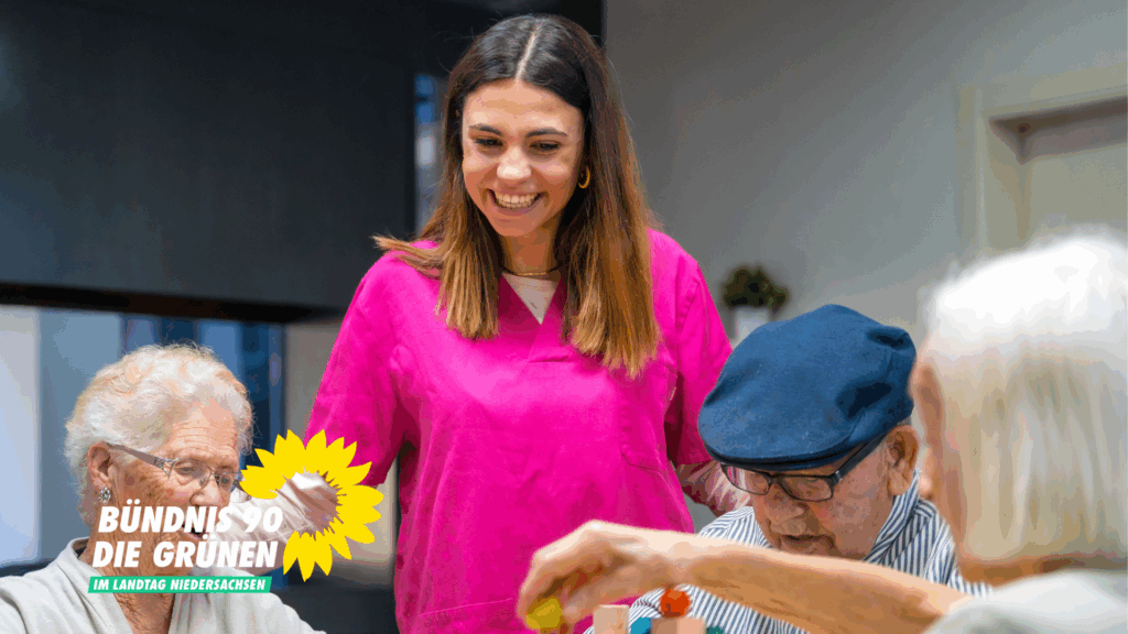 Drei ältere Personen sitzen an einem Tisch und spielen gemeinsam. Eine Pflegekraft im pinken Oberteil sieht ihnen dabei amüsiert zu.