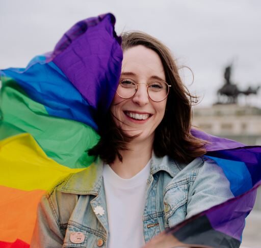 Swantje Schendel hat eine flatternde Regenbogenfahne um ihre Schultern gelegt und sieht dich fröhlich an.