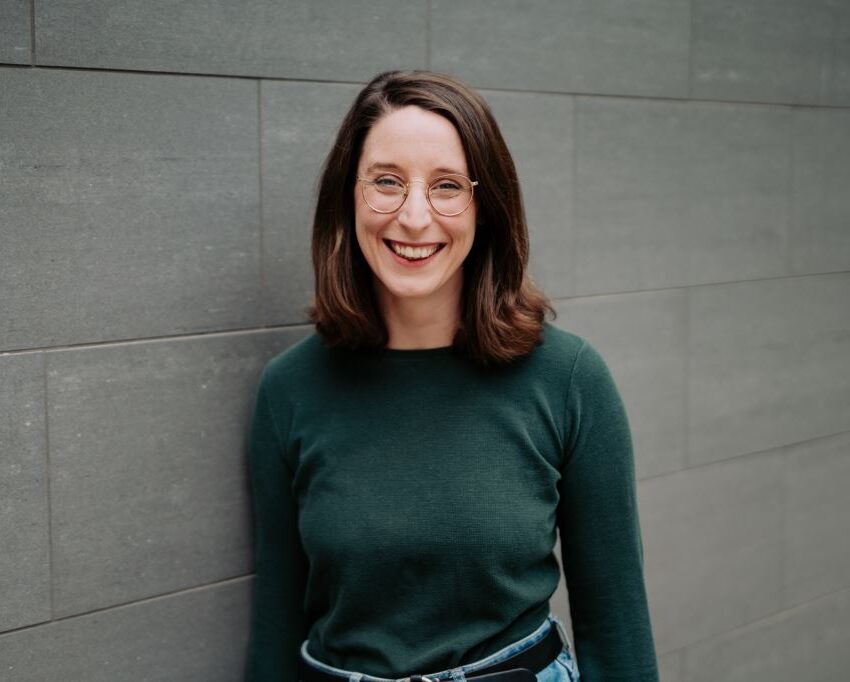 Swantje Schendel im dunkelgrünen Pullover steht vor einer grauen Wand und strahlt dich an.