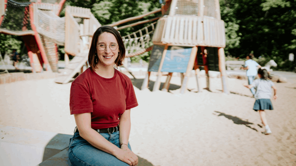 Swantje Schendel sitzt auf einem Spielplatz, im Hintergrund laufen Kinder durch den Sand auf ein Klettergerüst zu.