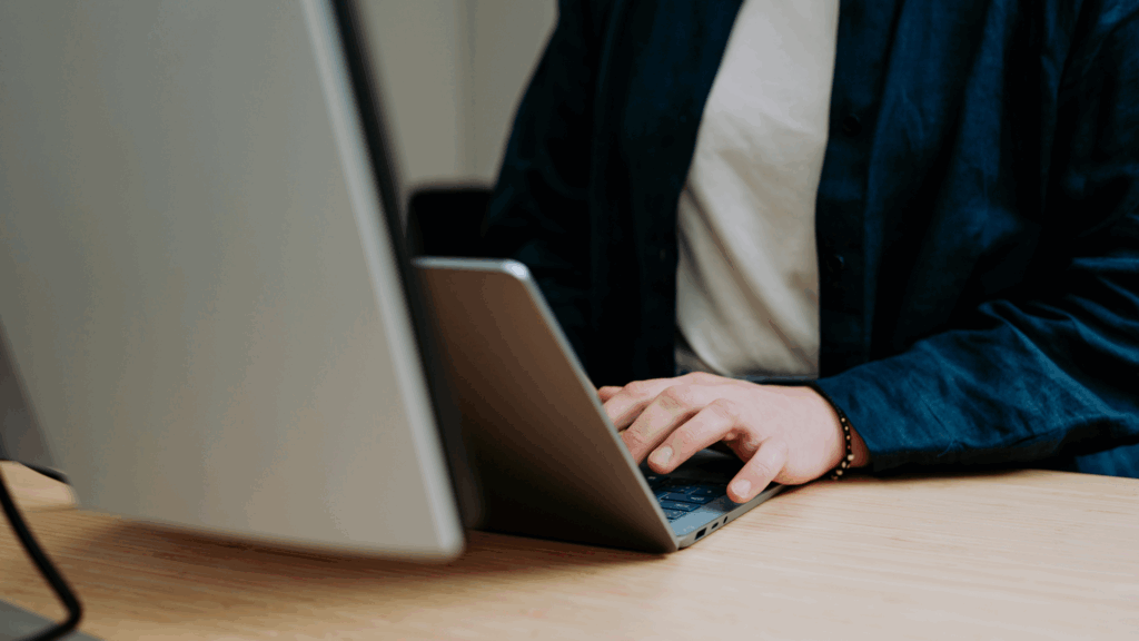 Zwei Hände tippen auf die Tastatur einer Laptops, davor ein großer Bildschirm.