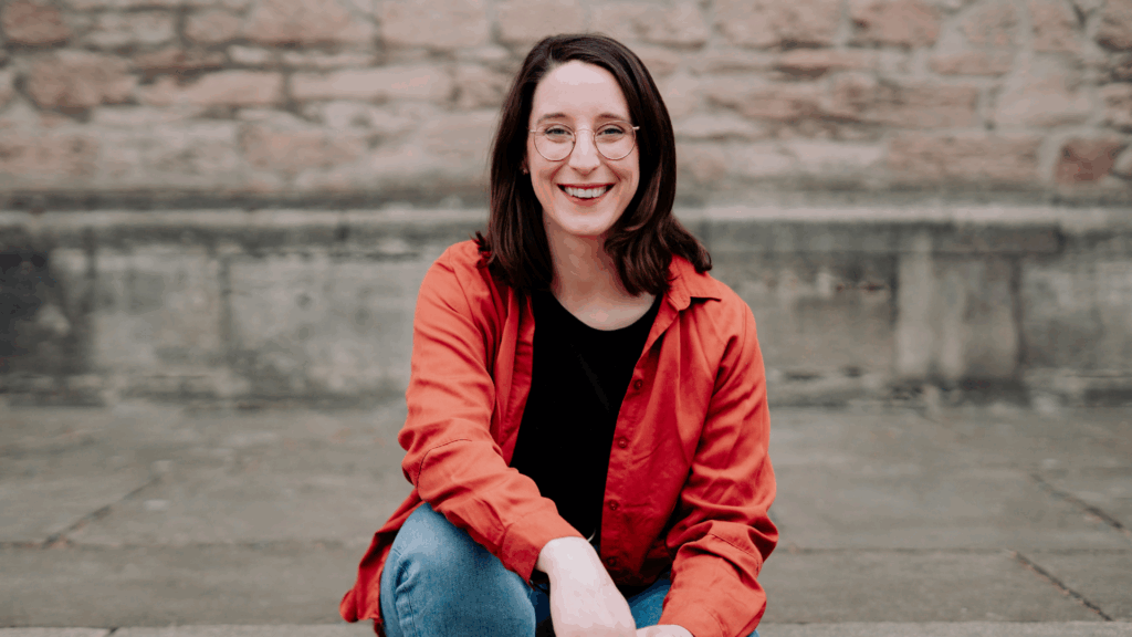 Swantje Schendel sitzt auf einem Bordstein und lächelt dich selbstbewusst an.